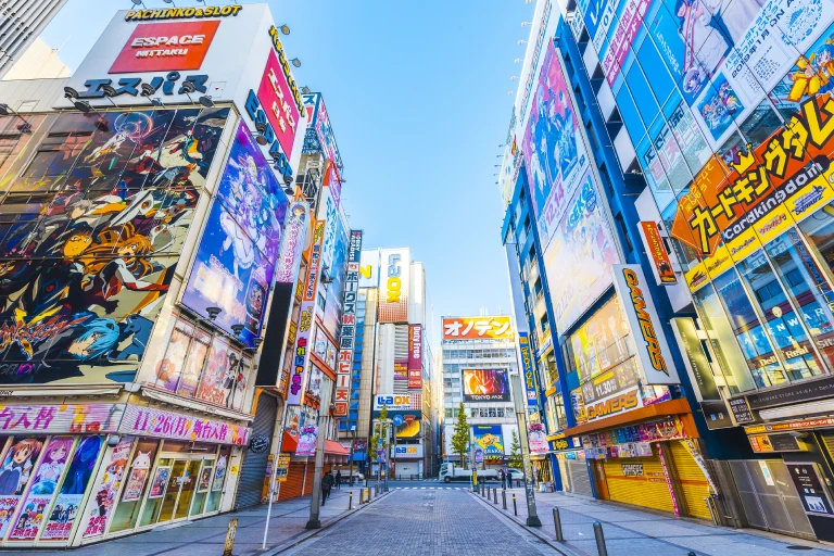 A bustling street in Akihabara, Tokyo, lined with colorful anime and gaming advertisements on tall buildings.