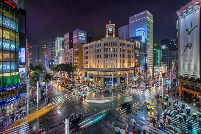 Night view of Ginza, Tokyo, Japan with illuminated buildings, busy streets, and people with umbrellas.