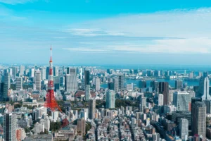 Aerial view of Tokyo cityscape with Tokyo Tower prominently in the center, surrounded by modern skyscrapers and buildings, with Tokyo Bay in the background under a clear blue sky.