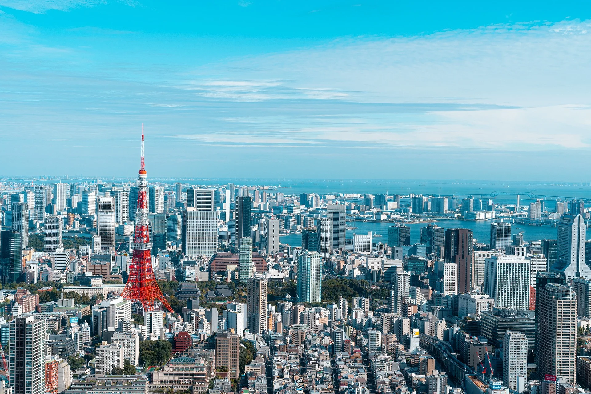 Aerial view of Tokyo cityscape with Tokyo Tower prominently in the center, surrounded by modern skyscrapers and buildings, with Tokyo Bay in the background under a clear blue sky.